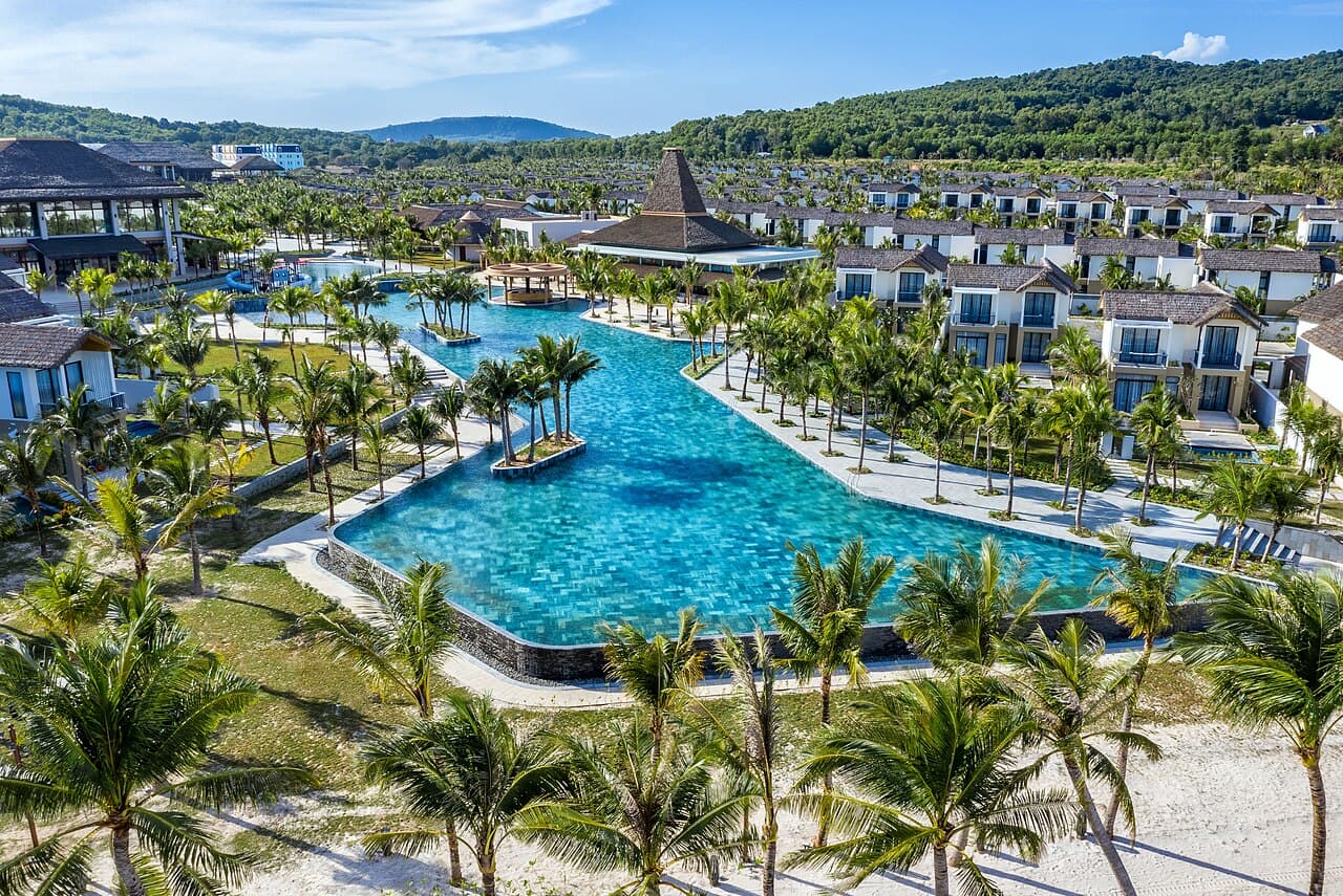 Các căn biệt thự hai tầng quanh một hồ bơi lớn giữa hàng dừa và thảm cỏ, nhìn từ trên cao