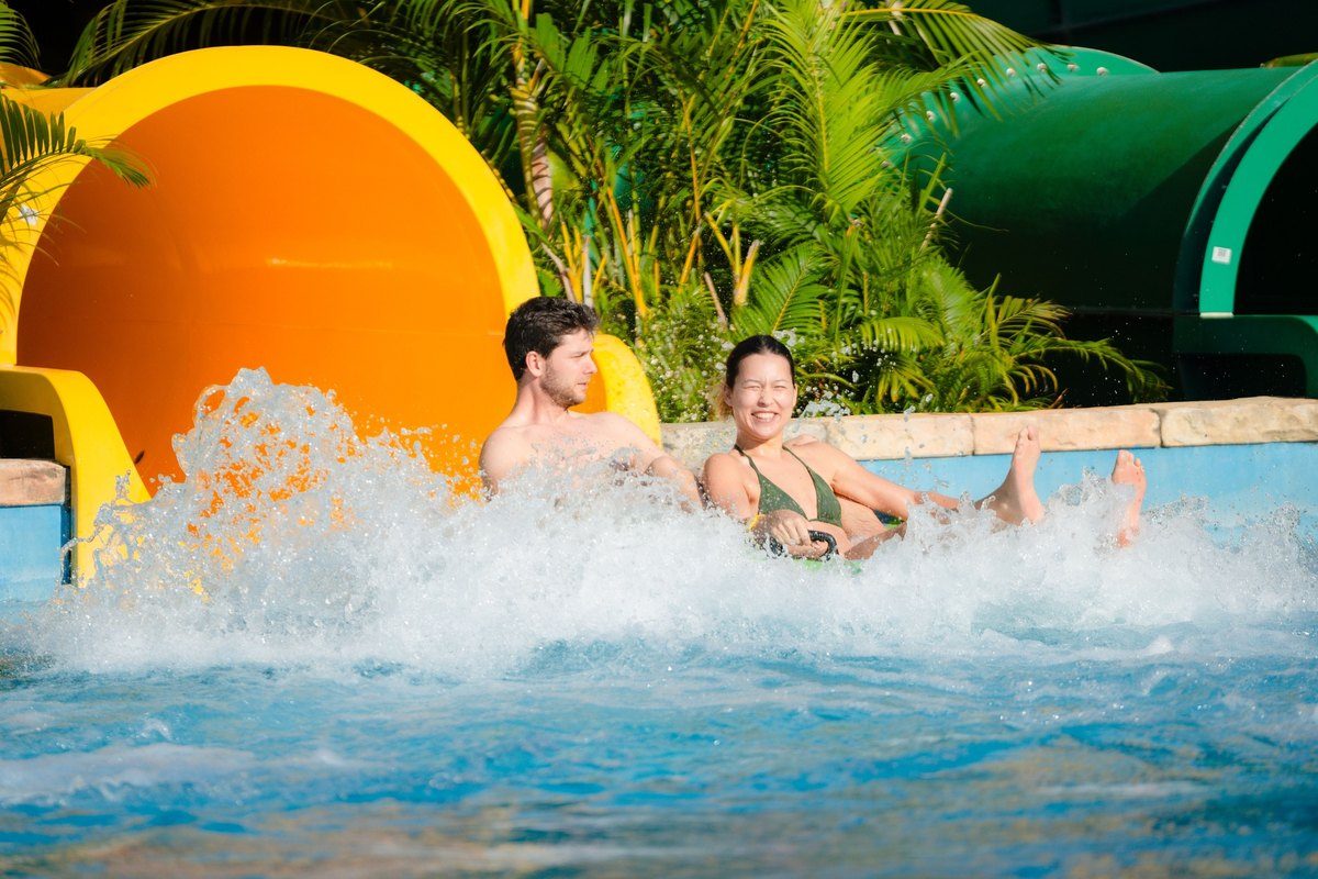Hai người lớn ngồi trên phao trượt ra khỏi ống trượt màu cam, nước bắn tung trắng xóa