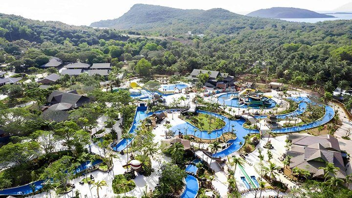 Nhìn từ trên cao các dòng nước và hồ bơi màu xanh uốn lượn giữa cây xanh và những mái nhà dốc, phía sau là núi