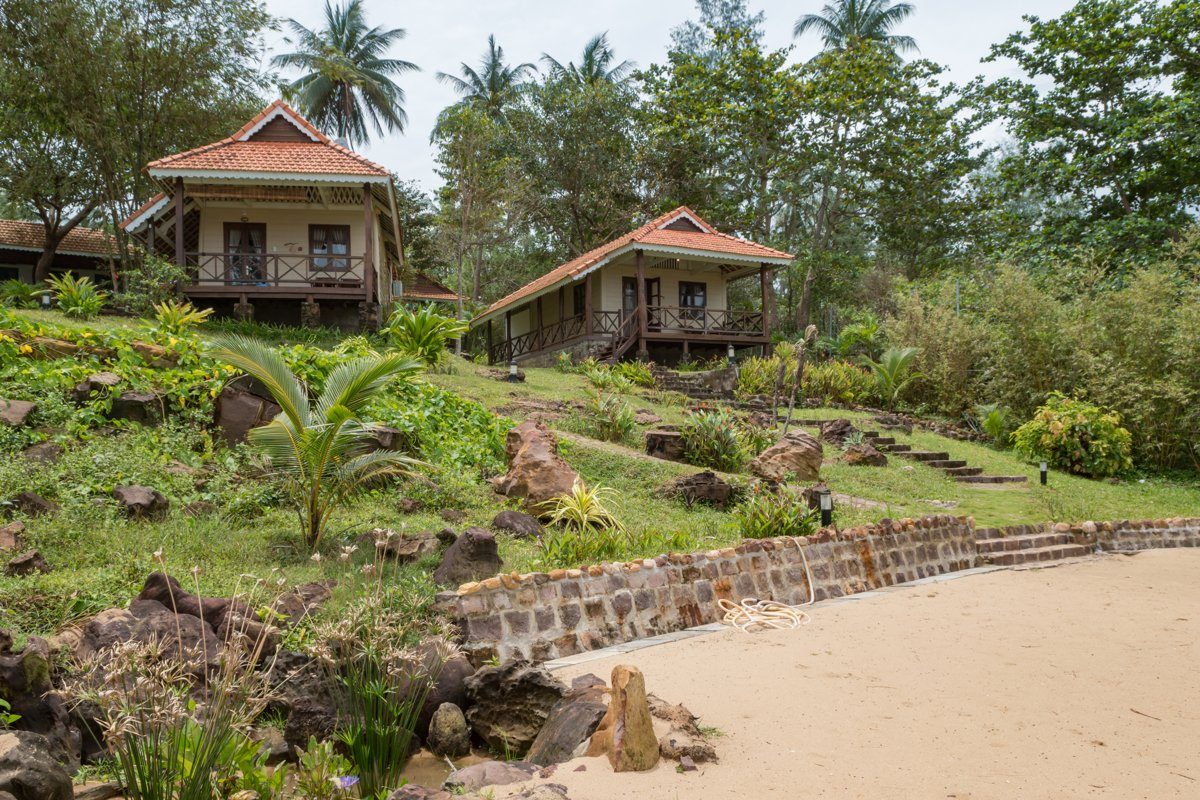 Hai căn bungalow gỗ mái ngói đỏ trên sườn dốc nhiều đá và cây xanh, phía dưới là bờ kè đá và bãi cát