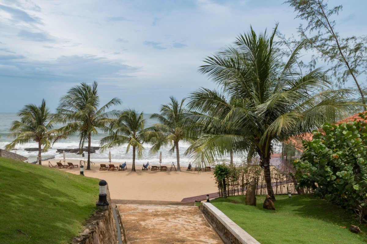Lối đi lát xuống dốc giữa hai bãi cỏ, dẫn ra bãi cát có hàng dừa và ghế dài, phía sau là biển và trời nhiều mây