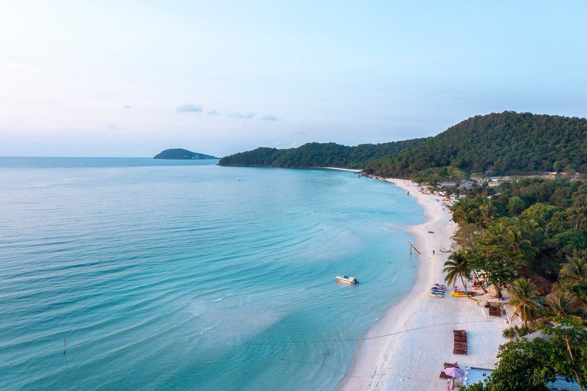 Bãi biển cát trắng cong dài bên làn nước xanh ngọc, hàng dừa và ghế nằm ven bờ, đồi rừng xanh và một hòn đảo nhỏ ở đường chân trời