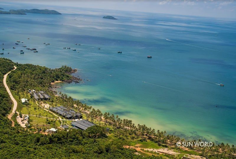 Bờ biển nhìn từ trên cao với rặng dừa dài, vài dãy mái nhà giữa cây xanh, nước nông xanh ngọc và nhiều thuyền nhỏ neo ngoài biển