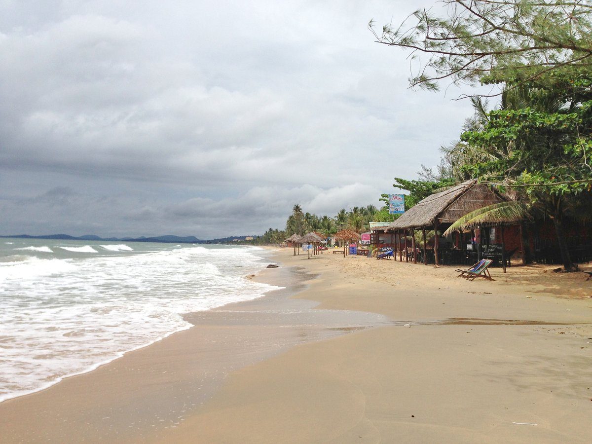 Bãi cát dài vắng người, sóng trắng dọc mép nước, bên phải là chòi lá và hàng cây, trời nhiều mây.