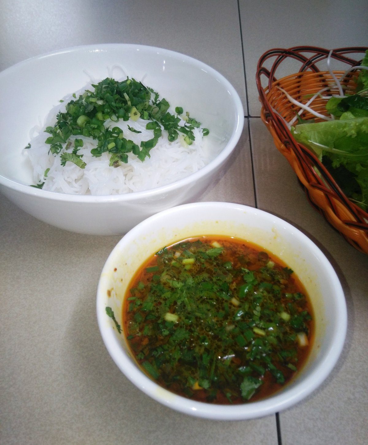 Tô bún trắng rắc hành lá và ngò, chén nước chấm đỏ cam đầy hành ngò, rổ rau sống đặt cạnh
