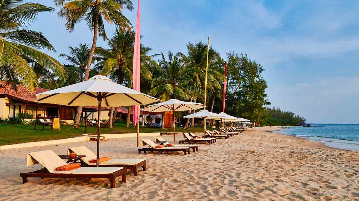 Bãi cát vàng với hàng ghế nằm gỗ và dù trắng, dừa và biệt thự phía sau, biển xanh bên phải.