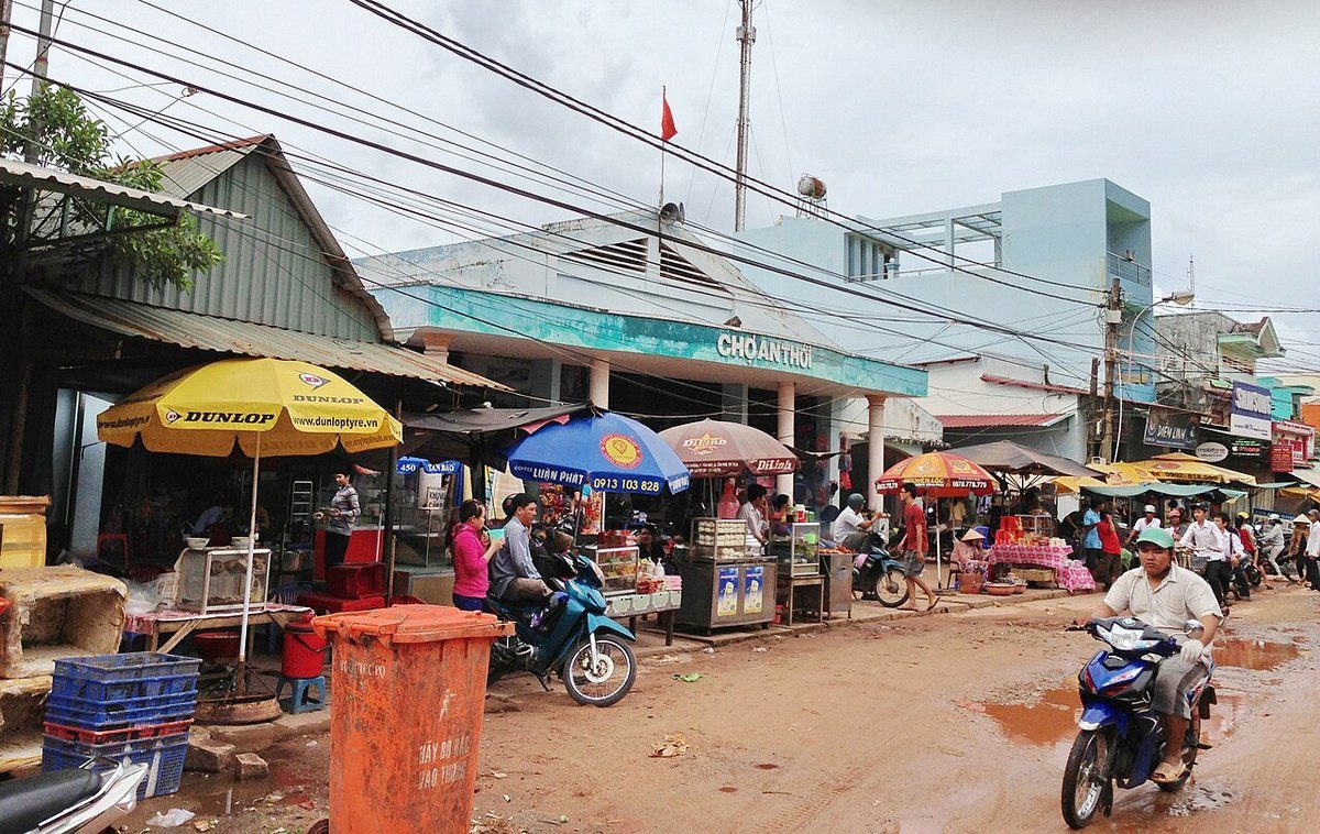 Mặt tiền chợ An Thới sơn xanh có chữ Chợ An Thới, phía trước là dù che, quầy hàng và người đi xe máy