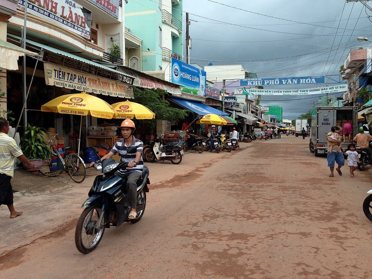 Đường phố ở khu chợ An Thới với tiệm tạp hóa, dù vàng, người đi xe máy và băng rôn căng ngang đường