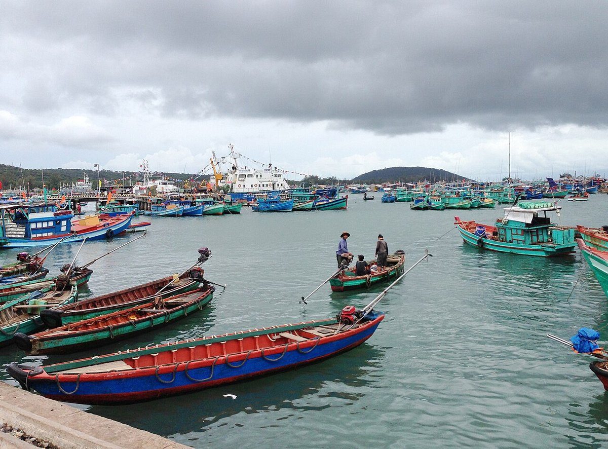 Những chiếc xuồng gỗ sơn đỏ và xanh neo sát bờ kè, phía sau là nhiều tàu cá dưới trời nhiều mây