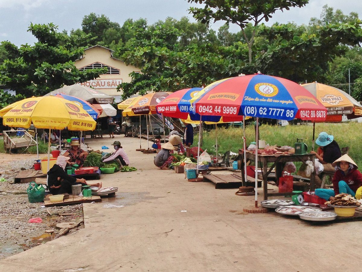 Người bán đội nón ngồi dưới dù màu bên mâm cá và cân, phía sau là nhà chợ sơn vàng có chữ Chợ Cầu Sấu