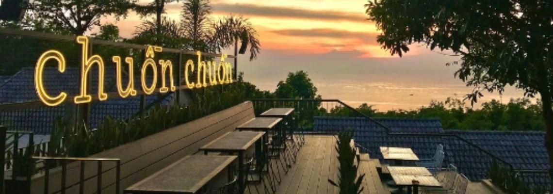 Chữ neon vàng chuồn chuồn trên lan can sân gỗ có dãy bàn, phía xa là mặt biển và bầu trời hoàng hôn màu cam