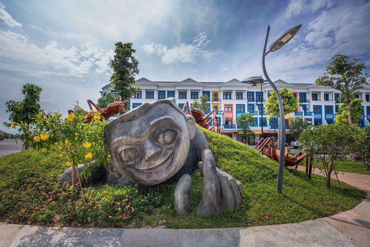 Tượng đá hình đầu người khổng lồ với đôi bàn tay nhô lên từ một gò cỏ, phía sau là tượng côn trùng và dãy nhà phố sơn nhiều màu