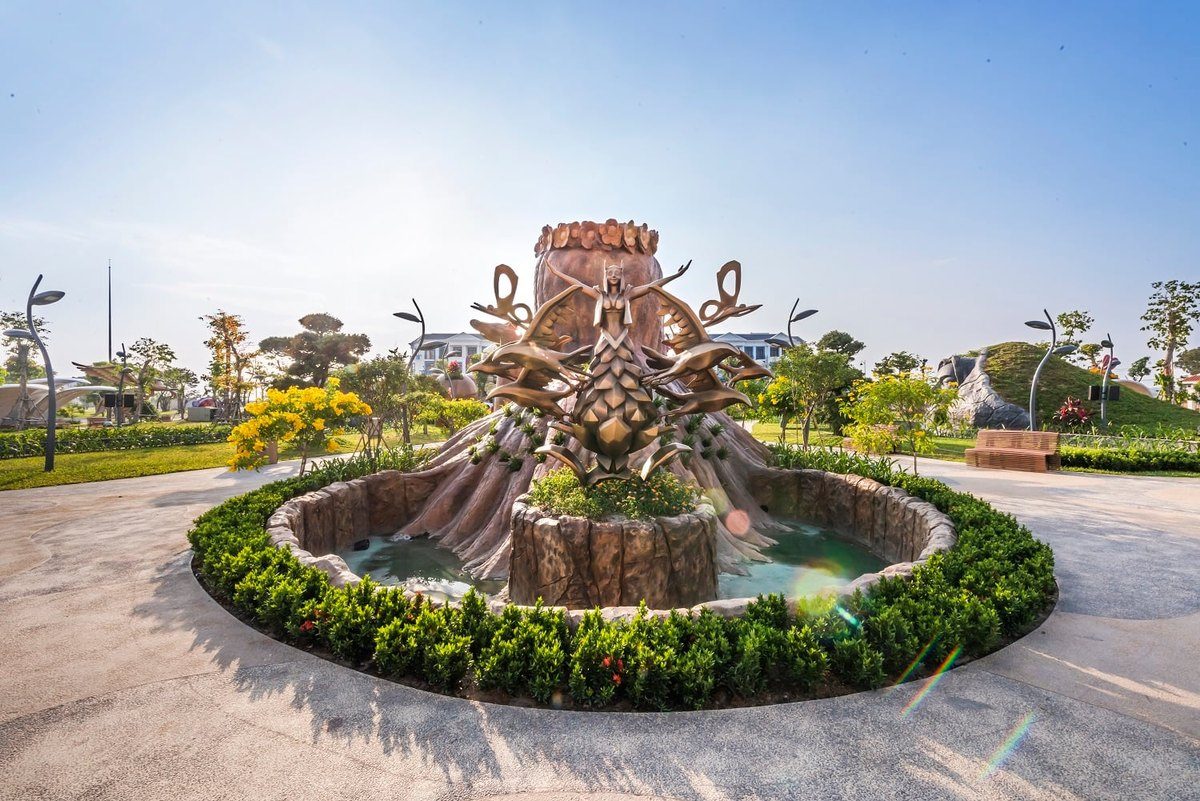Đài phun nước hình gốc cây lớn với nhiều hình người và nhánh cây bằng kim loại vươn ra, xung quanh là bồn hoa và lối đi lát gạch