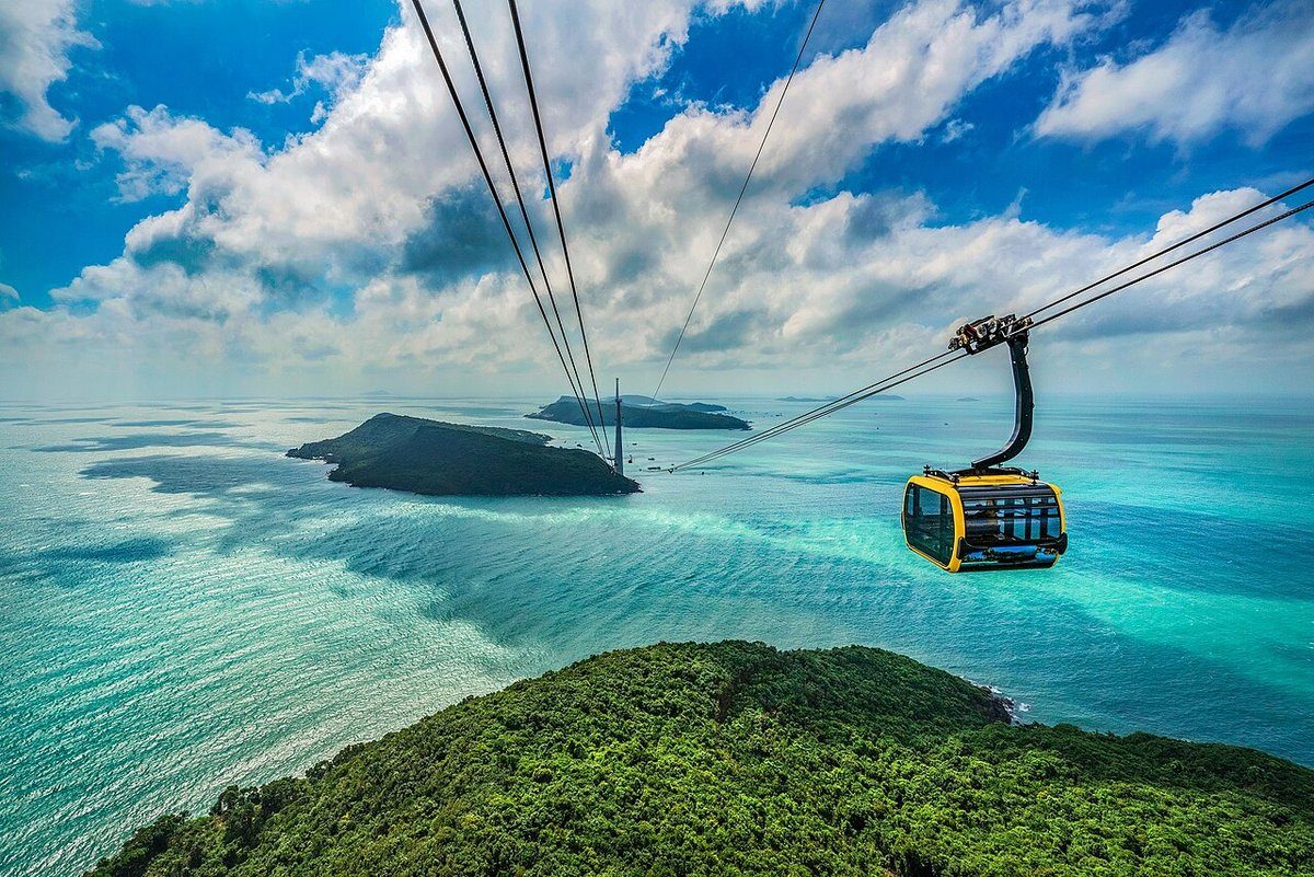Cabin cáp treo màu vàng treo trên ba dây cáp, bên dưới là đồi rừng xanh và biển xanh ngọc với các hòn đảo nhỏ