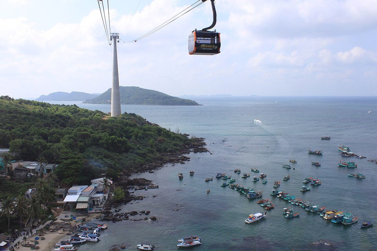 Cabin cáp treo trên cao, phía dưới là bờ biển An Thới với nhiều thuyền đánh cá neo trên mặt nước, xa xa là trụ cáp và hòn đảo