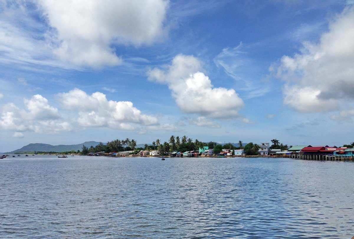 Dãy nhà ven biển của làng chài Hàm Ninh nhìn từ mặt nước, phía xa là núi, trời nhiều mây.