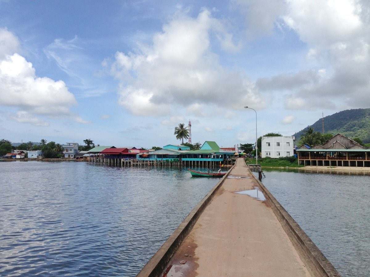Cầu bê tông dẫn vào bờ, cuối cầu là dãy nhà hàng dựng trên cọc và núi xanh.