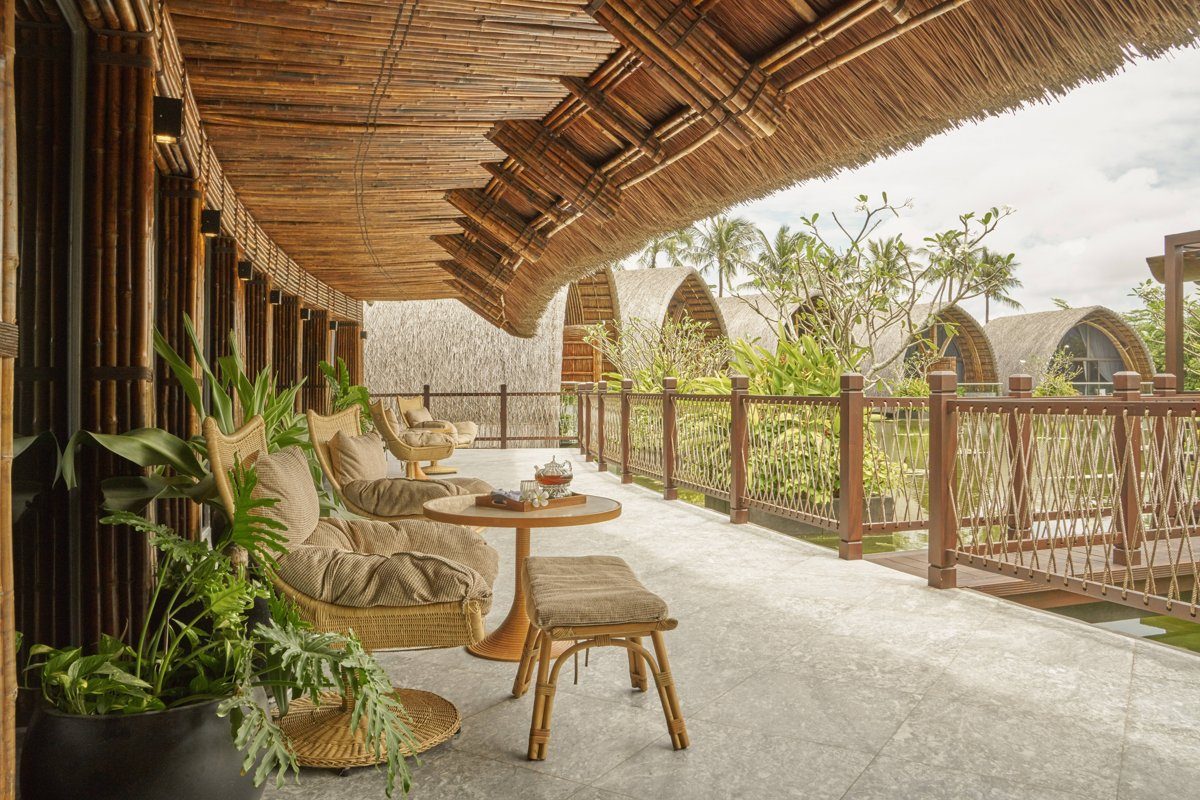 Hiên gỗ có ghế mây và bàn trà dưới mái tre, nhìn ra những mái spa cong giữa cây xanh