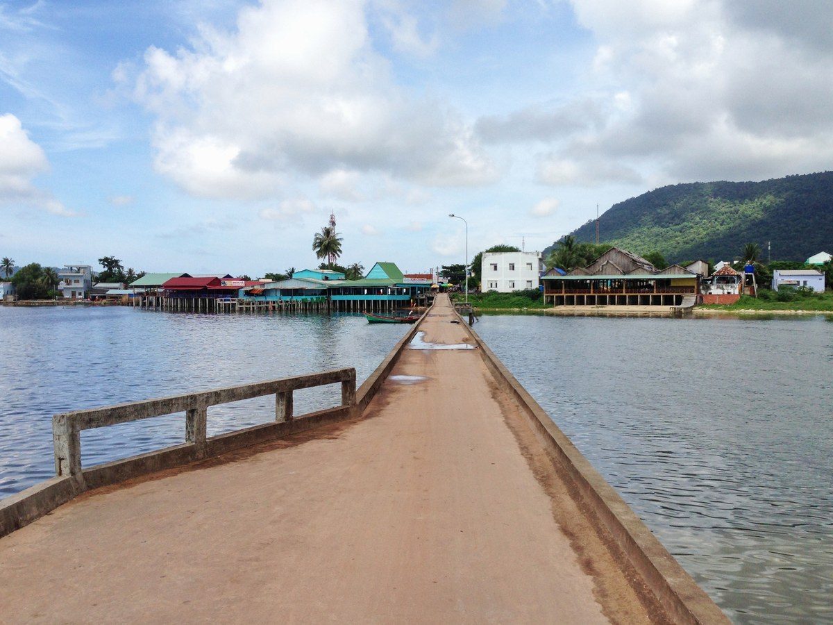 Cầu bê tông hẹp chạy vào bờ, cuối cầu là dãy nhà mái đỏ, mái xanh dựng trên cọc và núi xanh phía sau.