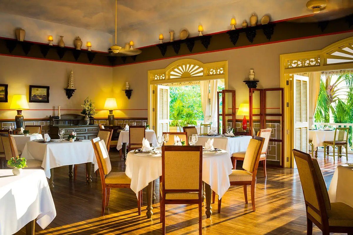 Phòng ăn trải khăn bàn trắng, trần cao, cửa vòm kiểu thuộc địa mở ra khu vườn nhiệt đới.