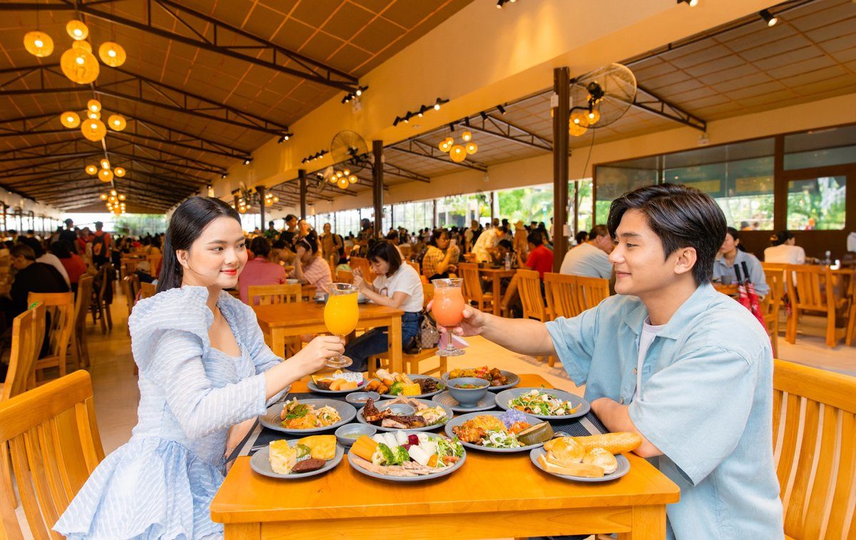 Sảnh nhà hàng buffet Mango với mái cao, đèn treo và nhiều dãy bàn gỗ, hai người nâng ly ở bàn phía trước