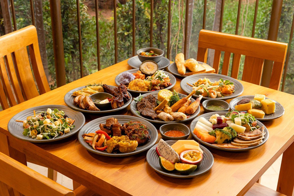 Bàn gỗ bày nhiều đĩa thức ăn buffet gồm thịt, hải sản, salad, bánh mì và trái cây