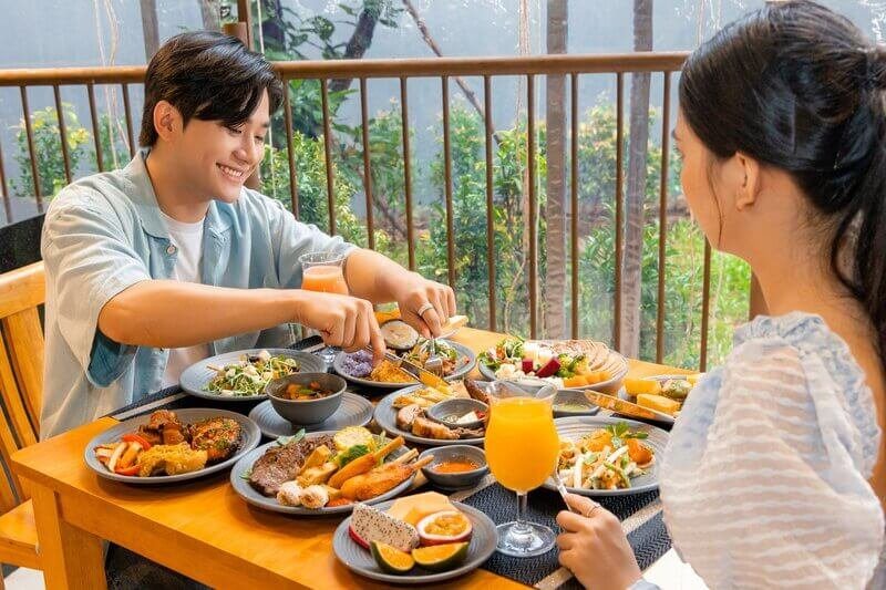 Hai người ngồi ăn trưa ở bàn gỗ cạnh lan can, trên bàn có nhiều đĩa thức ăn và ly nước ép cam