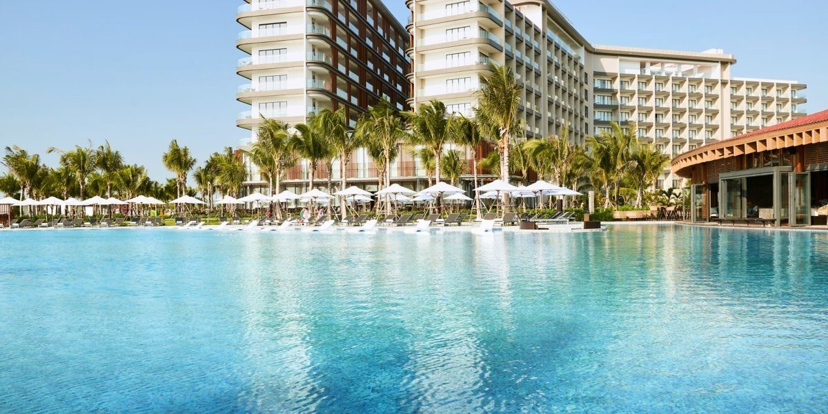 Hồ bơi xanh trong phía trước, hàng dừa và ô trắng, hai tòa nhà khách sạn cao tầng phía sau.