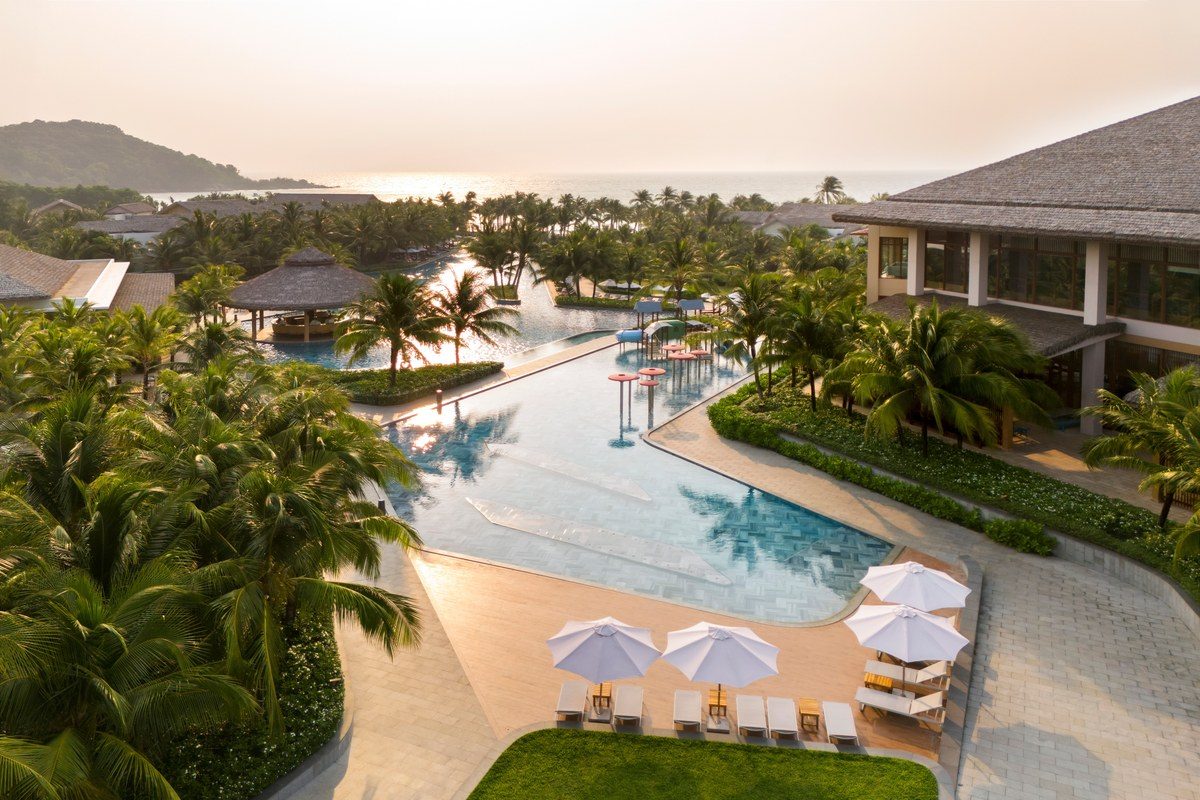 Hồ bơi dài uốn lượn giữa hàng dừa và mái tranh, biển và núi phía sau, nhìn từ trên cao lúc chiều.