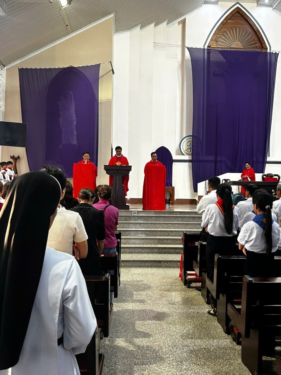 Bên trong nhà thờ trắng với vòm nhọn và màn tím, ba linh mục áo đỏ đứng trên bậc cung thánh, giáo dân đứng giữa các hàng ghế gỗ nhìn lên