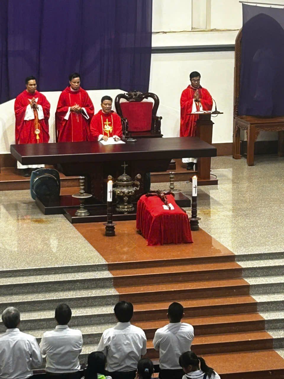 Bàn thờ gỗ sẫm màu trên cung thánh, phía trước là thánh giá đặt trên bục phủ vải đỏ giữa hai cây nến, bốn linh mục mặc áo lễ đỏ đứng phía sau