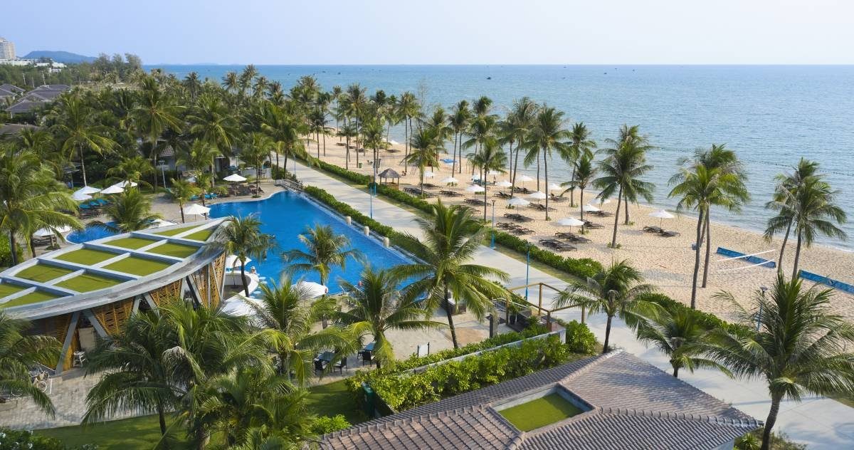 Toàn cảnh từ trên cao một hồ bơi dài màu xanh giữa hàng dừa, cạnh bãi cát có dù trắng và biển xanh