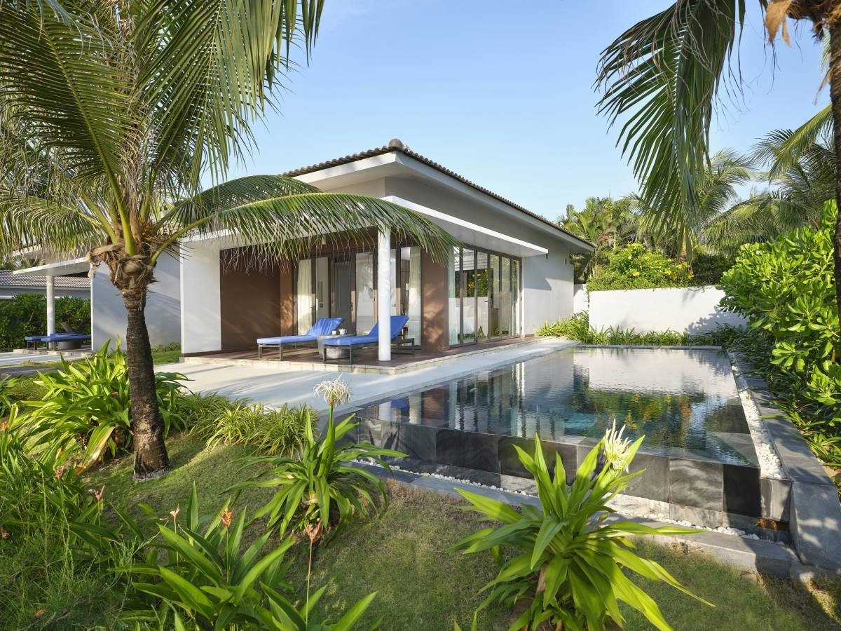 Một căn bungalow màu trắng có hiên rộng và hồ bơi riêng, xung quanh là cây dừa và cây bụi xanh