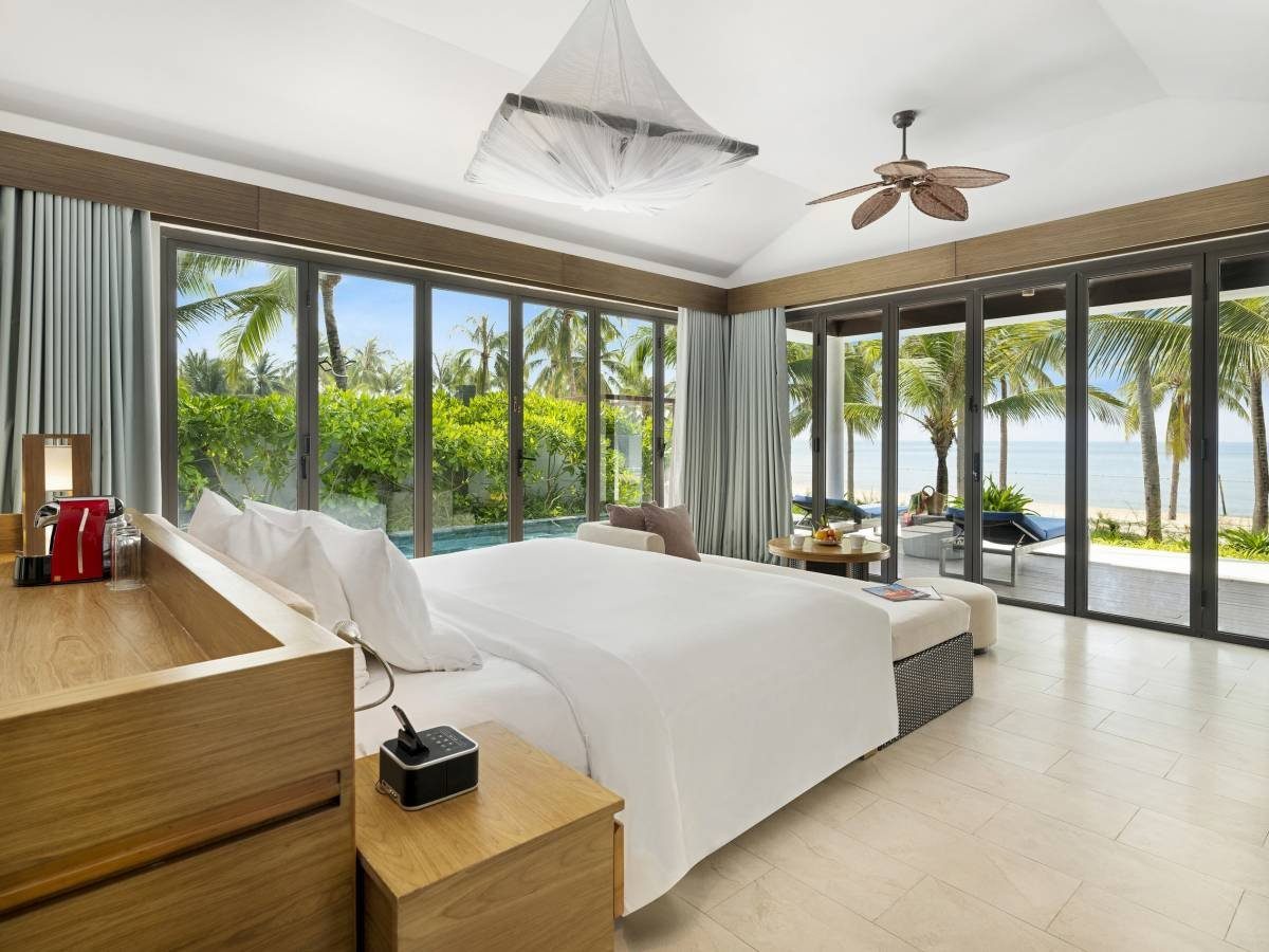 Phòng ngủ sáng với giường đôi trắng, quạt trần, cửa kính lớn nhìn ra hồ bơi riêng và biển