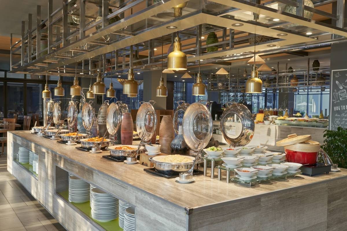 Quầy buffet dài bằng gỗ với nhiều nồi nắp bạc, đĩa thức ăn và đèn treo màu đồng phía trên