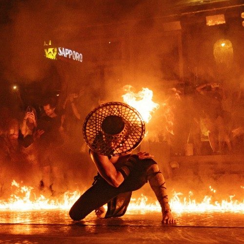 Người biểu diễn đội nón lưới quỳ giữa vòng lửa trên sàn, phía sau là khách đứng xem và quay điện thoại