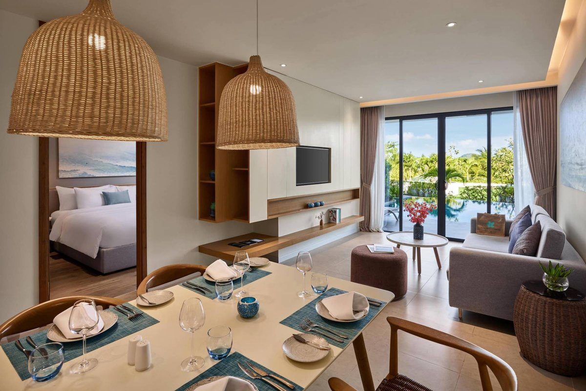 Phòng khách có bàn ăn, sofa, đèn mây đan, cửa kính lớn nhìn ra hồ bơi riêng và cây xanh