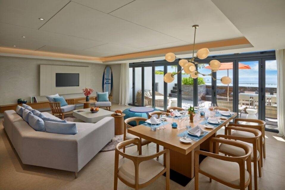 Phòng khách rộng có sofa và bàn ăn tám ghế, cửa kính mở ra sân hiên có dù và biển phía xa