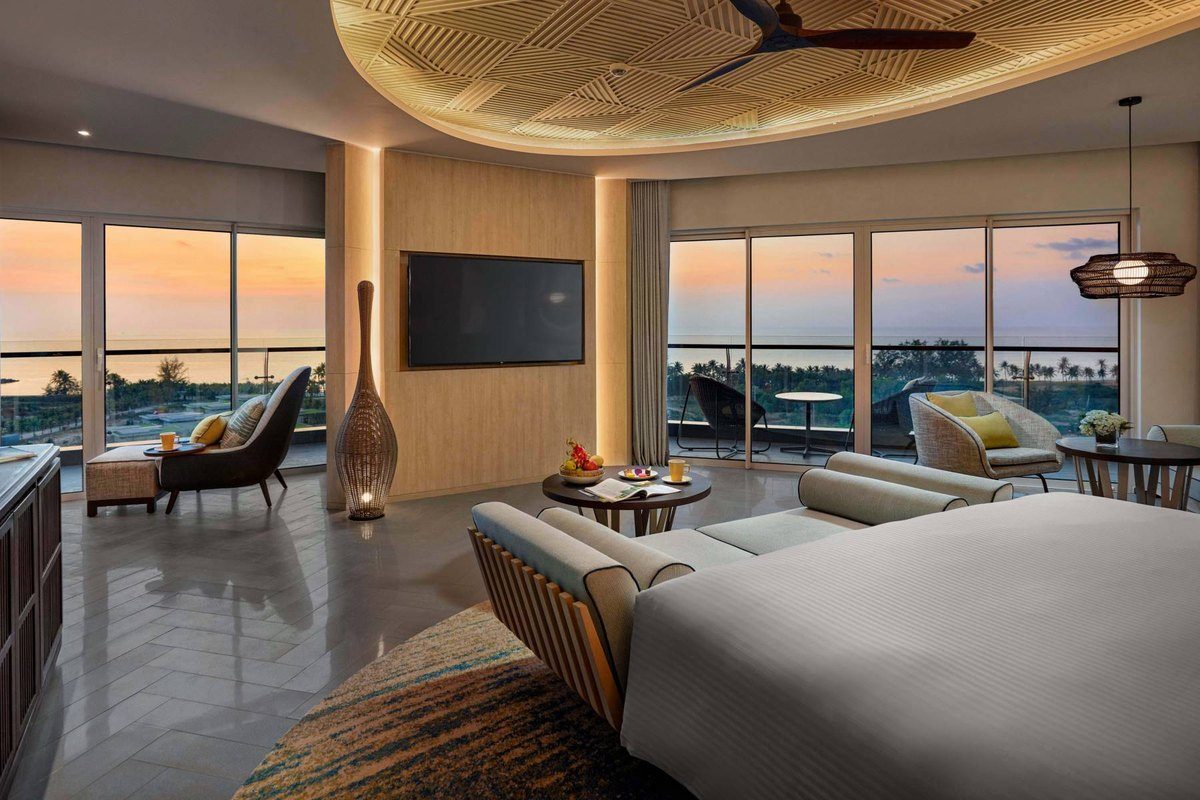 Phòng suite có giường lớn, sofa và cửa kính rộng nhìn ra biển lúc hoàng hôn