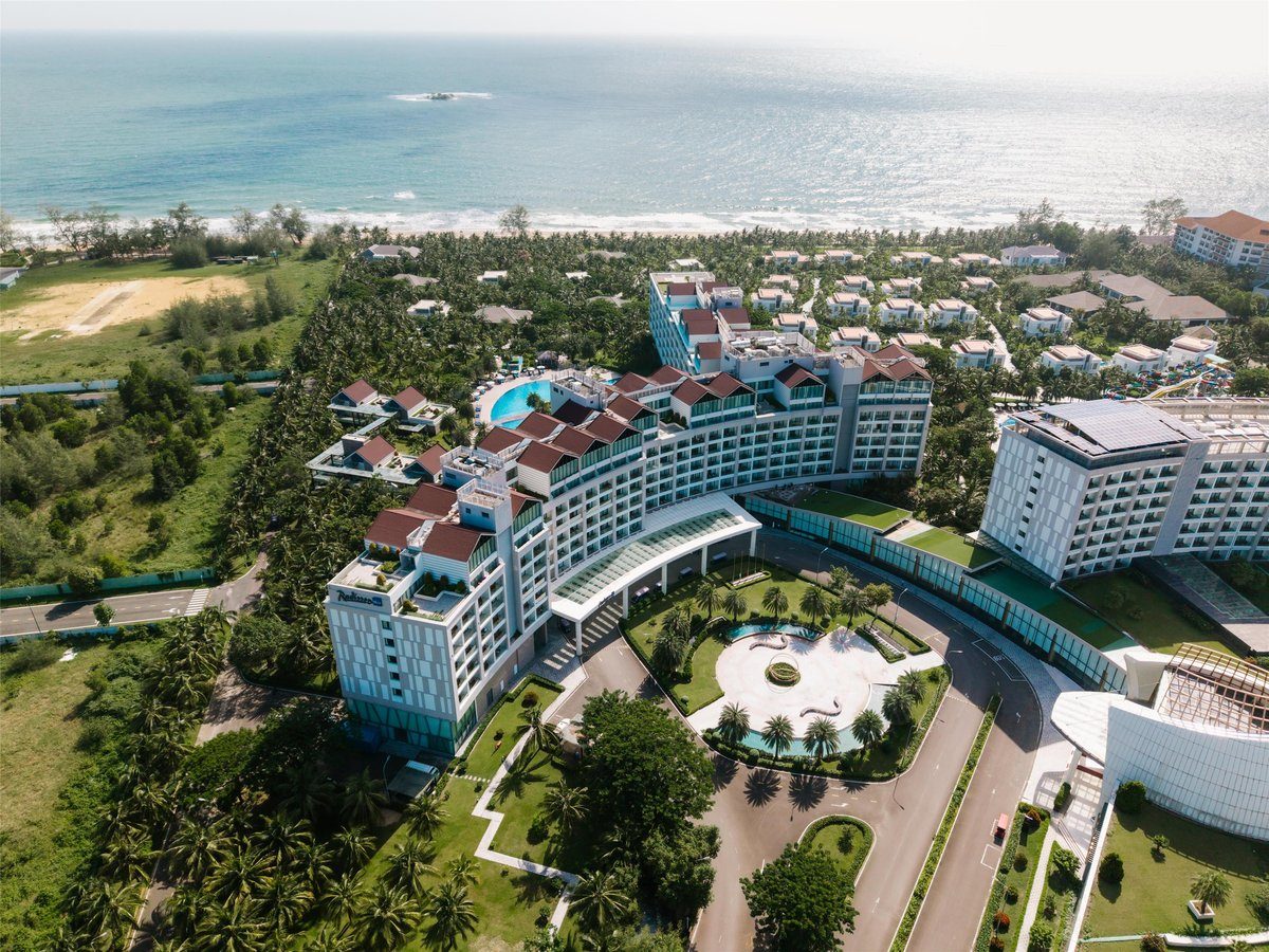 Ảnh chụp từ trên cao khu nghỉ dưỡng với các tòa nhà trắng mái đỏ, vườn cây và bờ biển phía sau.