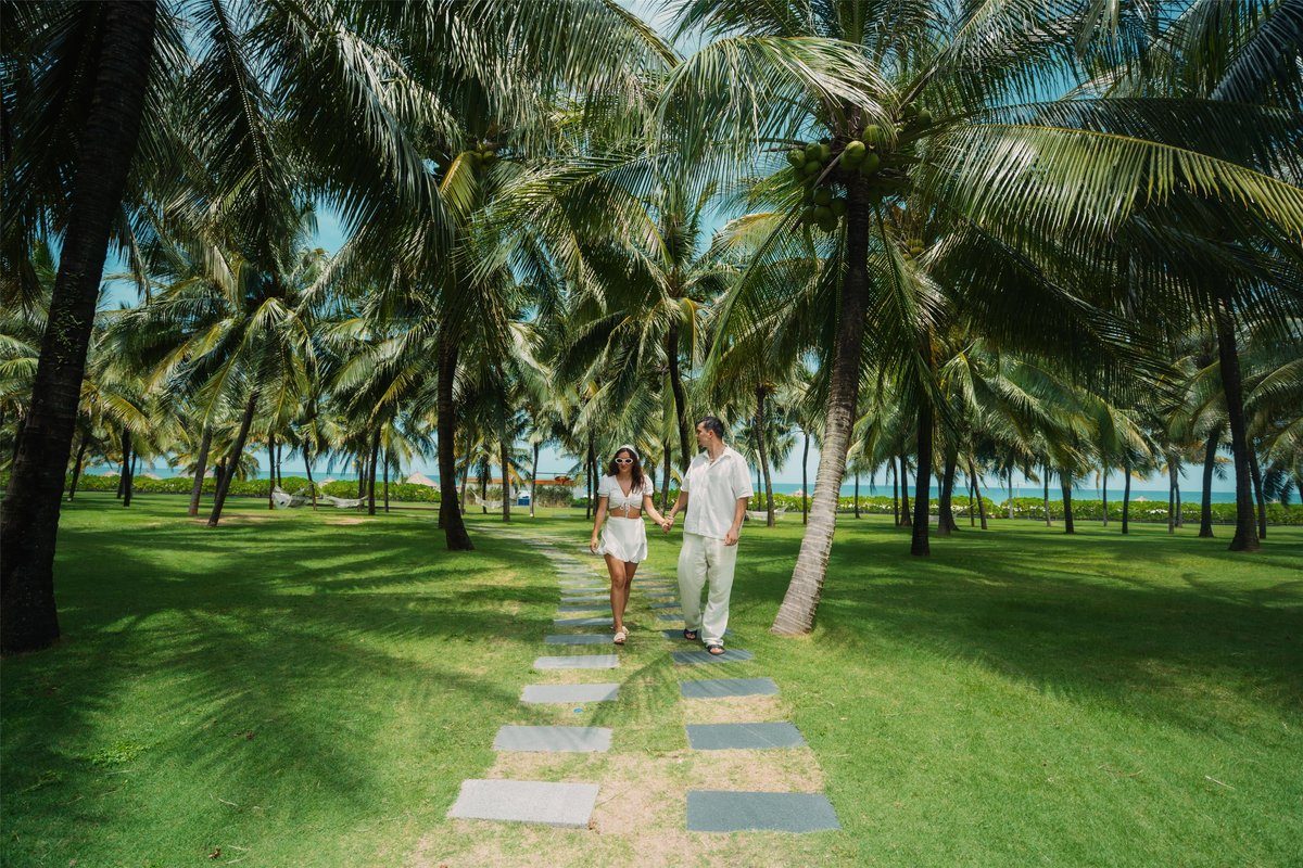 Hai người đi trên lối lát đá giữa bãi cỏ và hàng dừa, phía xa là biển.