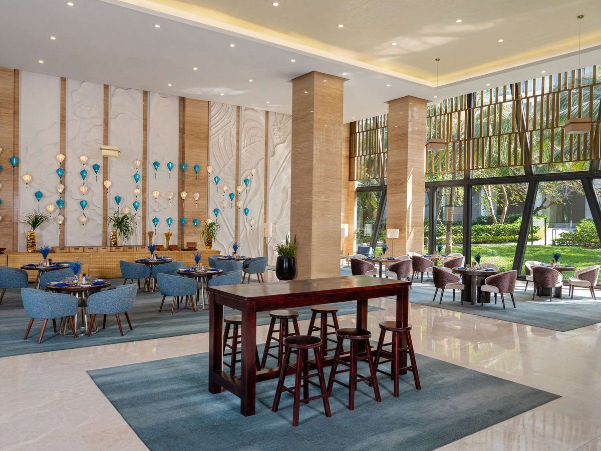 Không gian lounge trần cao với cột ốp gỗ, ghế bọc nệm xanh, bàn cao và vách kính nhìn ra vườn.