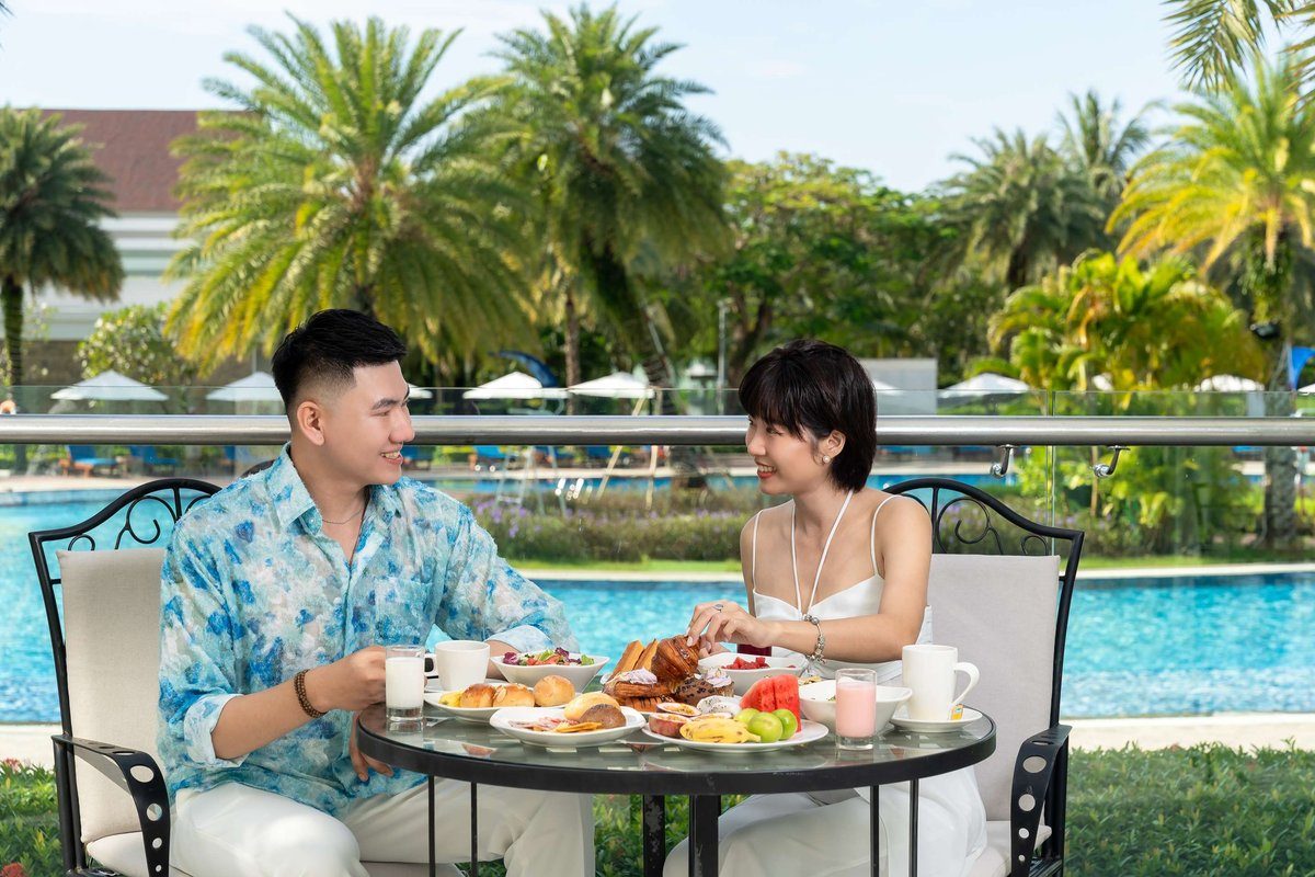 Hai người lớn ăn sáng ở bàn tròn ngoài trời, phía sau là hồ bơi và hàng cọ.
