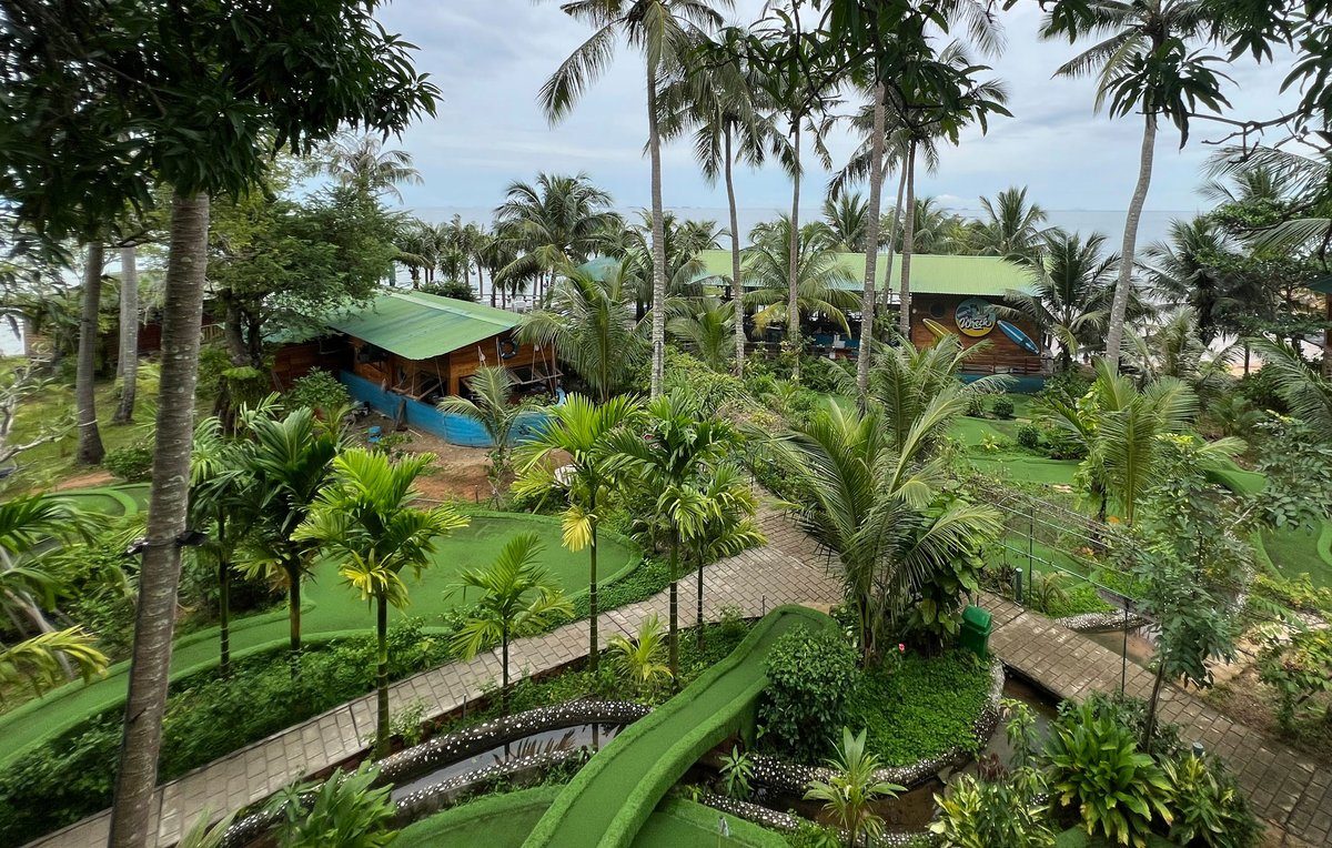 Khu vườn nhiệt đới nhìn từ trên cao, các đường cỏ nhân tạo xanh uốn lượn giữa cây dừa và lối đi, phía sau là những mái nhà màu xanh