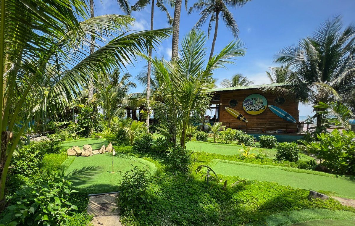 Thảm cỏ golf mini có cắm cờ, vài tảng đá, cây dừa xung quanh, phía sau là căn nhà gỗ có biển hiệu tròn và hai tấm ván lướt sóng