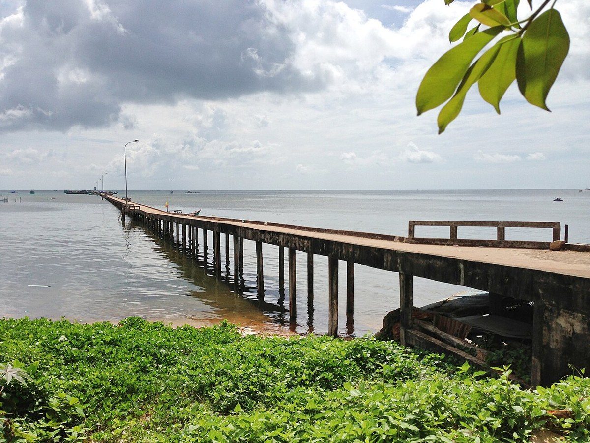 Cầu tàu bê tông trên hàng cọc chạy ra mặt biển lặng, phía trước là bụi cây xanh trên bờ