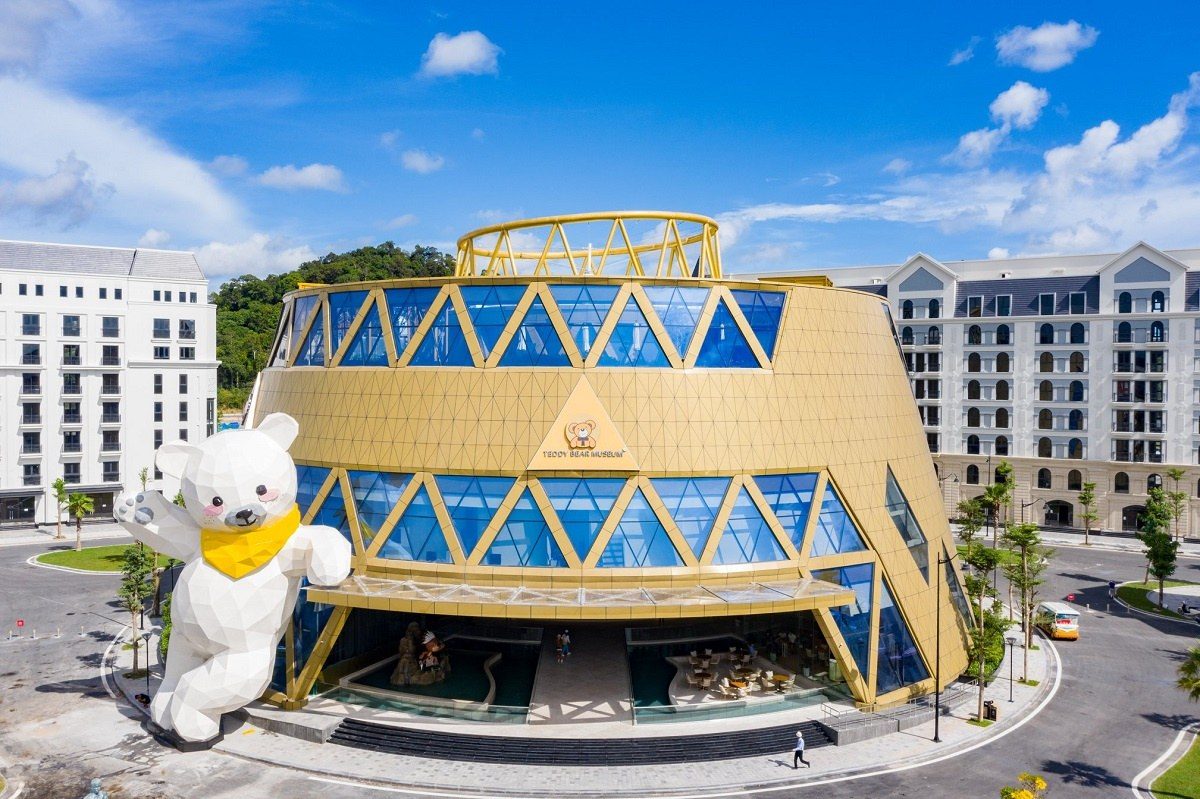 Tòa nhà hình tròn ốp tấm vàng và kính xanh, biển Teddy Bear Museum ở giữa, bên trái là tượng gấu trắng khổng lồ đeo khăn vàng, hai bên là dãy nhà nhiều tầng