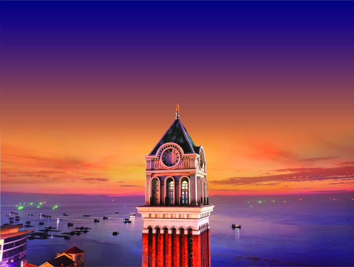 Phần đỉnh tháp đồng hồ với mái chóp nhọn màu xanh, mặt đồng hồ tròn và các cửa vòm, phía sau là bầu trời cam tím và mặt biển có thuyền sáng đèn