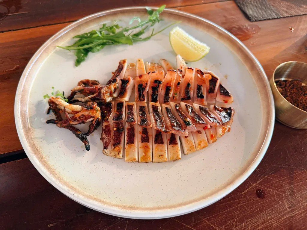 Con mực nướng nguyên con khứa ô vuông trên đĩa gốm trắng, kèm lát chanh và rau xanh, đặt trên bàn gỗ