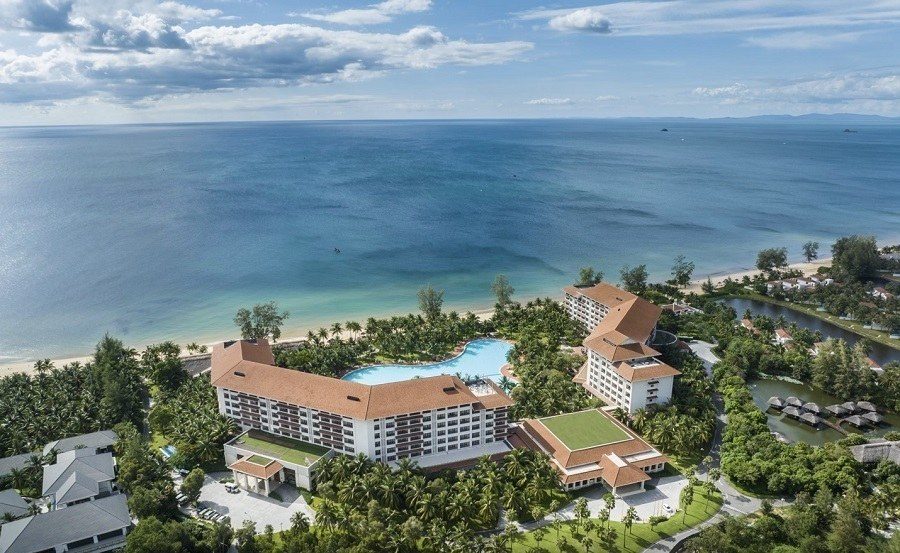 Ảnh chụp từ trên cao khu nghỉ với các tòa nhà mái ngói đỏ, hồ bơi xanh giữa vườn cây, bãi cát và biển phía sau.